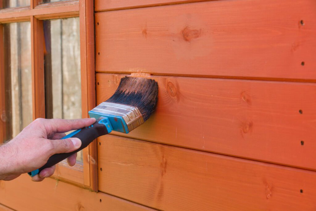 A person using a paint brush to paint a shed orange.